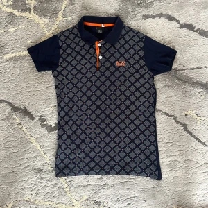 Mörkblå pikétröja från BOSS - Snygg mörkblå pikétröja från BOSS med korta ärmar och krage. Tröjan har ett rutigt mönster framtill, orange detaljer vid knapparna och BOSS-logga broderad på bröstet. Perfekt för en stilren look.