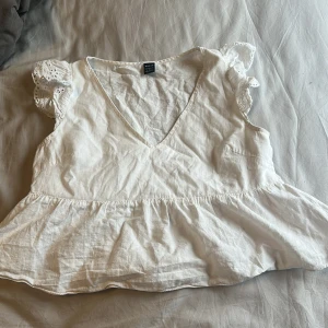 Vit blus med spetsdetaljer från SHEIN - Säljer en söt vit blus från SHEIN med v-ringning och volang nertill. Blusen har korta ärmar med broderad spets och blommiga detaljer. Perfekt för vår och sommar!