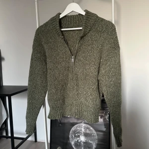 Olivgrön stickad hoodie med dragkedja - Mysig olivgrön stickad hoodie med halv dragkedja framtill och ribbade muddar. Tröjan är tillverkad i 100% polyester och har en avslappnad passform med lång ärm. Perfekt för kyligare dagar.