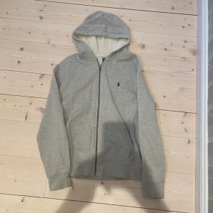 Grå hoodie från Polo Ralph Lauren - Säljer en klassisk grå hoodie från Polo Ralph Lauren med dragkedja och huva. Tröjan har det ikoniska broderade logotypen på bröstet och är tillverkad i mjuk bomullsmix. Perfekt för en avslappnad stil. Storlek XL men passar även L. Skick 7/10, skriv om ni undrar något!