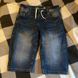 Blå jeansshorts från LAB Industries - Snygga blå jeansshorts från LAB Industries med vit snörning i midjan och klassiska fem fickor. Shortsens denim har en lätt tvättad look och de går ner till knäna. Perfekta för en avslappnad stil under varmare dagar.