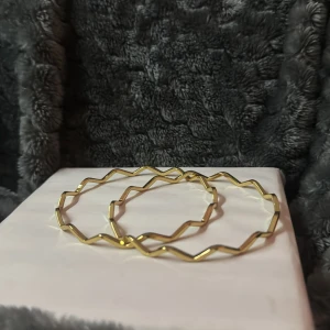 Guldiga zickzack-formade armband  - Säljer ett par stora, runda armband i guld med unik zickzack-form. De är tunna och har en modern, geometrisk design som sticker ut. Perfekta för dig som vill ha något annorlunda och trendigt i smyckeskrinet.