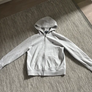Grå hoodie från Polo Ralph Lauren - Säljer en stilren grå hoodie från Polo Ralph Lauren med dragkedja och huva. Tröjan har långa ärmar, två fickor framtill och den klassiska broderade loggan på bröstet. Den är i ett väldigt bra skick så bara höra av er om ni vill ha fler bilder eller om det är något ni undrar över
