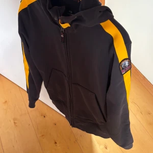 Svart hoodiejacka med gula ränder från Parajumpers - Svart hoodiejacka med dragkedja och huva från Parajumpers. Gula ränder längs ärmarna och logotyp på vänster ärm. Två fickor framtill och ribbade muddar. Perfekt för en sportig stil.