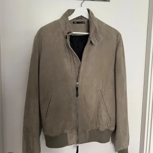 Beige mockajacka från Zara - Snygg beige mockajacka från Zara med dragkedja framtill och ribbade muddar vid ärmslut och midja. Jackan har en klassisk krage och två sidofickor. Perfekt för dig som vill ha en stilren och tidlös look.