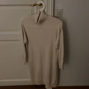 Beige stickad poloklänning - Mysig beige stickad klänning med polokrage och långa ärmar. Klänningen har ribbade muddar vid ärmslut och nederkant, vilket ger en snygg och bekväm passform. Perfekt för kyligare dagar och enkel att styla med boots eller sneakers.