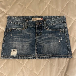 Blå jeanskjol  - Säljer en blå jeanskjol i klassisk denim med slitna detaljer och fickor både fram och bak. Kjolen har bälteshällor och knappstängning framtill. Perfekt för dig som gillar en tuff och avslappnad stil. Den är lågmidjade, BARA INTRESSEKOL!!