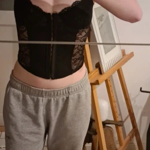 Svart spetskorsett med dragkedja - Säljer en svart korsett med spetsdetaljer och dragkedja framtill. Toppen har smala axelband och är genombruten med blommönstrad spets på sidorna. Perfekt för att skapa en snygg och trendig look.