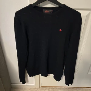 Marinblå stickad tröja från Morris - Säljer en marinblå stickad tröja från Morris med rund halsringning och diskret röd broderad logga på bröstet. Tröjan har lång ärm och klassisk passform, perfekt för att kunna matcha med allt