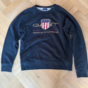 Svart sweatshirt från Gant med broderad logga - Svart sweatshirt från Gant med broderad GANT-logga och sköld på bröstet. Tröjan har rund halsringning, ribbade muddar och klassisk passform. Perfekt för en sportig och stilren look.