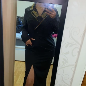 Svart långklänning med broderad krage - Säljer en svart långklänning med elegant broderad krage i guld. Klänningen har lång ärm och en snygg slits framtill. Perfekt för dig som vill sticka ut med stilrena detaljer och en klassisk siluett.