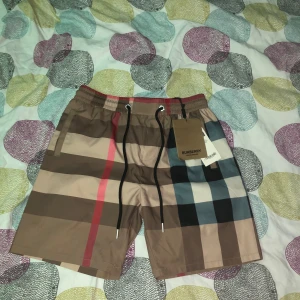 Rutiga badshorts från Burberry - Snygga badshorts från Burberry med klassiskt rutigt mönster i beige, svart, rött och blått. Shortsen har snörning i midjan och sidofickor. Perfekta för sommaren och strandhäng.
