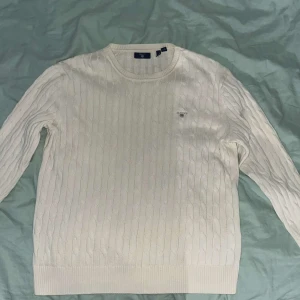 Vit kabelstickad tröja från GANT - Säljer en klassisk vit kabelstickad tröja från GANT med rund halsringning och diskret broderad logga på bröstet. Tröjan har långa ärmar och ribbade muddar vid ärmslut och nederkant. Perfekt för en stilren look.