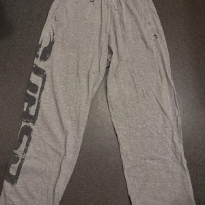 Grå Gasp byxor med tryck - Säljer ett par grå byxor från gasp med svart tryck längs ena benet. Byxorna har resår och snörning i midjan för extra komfort. Perfekta för chill eller träning. Mjukt material och avslappnad passform.