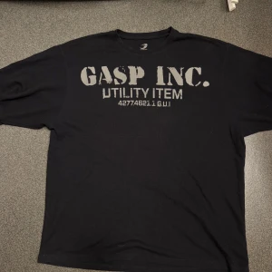 Svart t-shirt från GASP INC. - Svart t-shirt från GASP INC. med tryck både fram och bak. Framsidan har texten 'GASP INC. UTILITY ITEM' i grått och baksidan har ett stort 'GASP'-tryck. Klassisk rund hals och oversized passform. Perfekt för dig som gillar en bra pump cover.