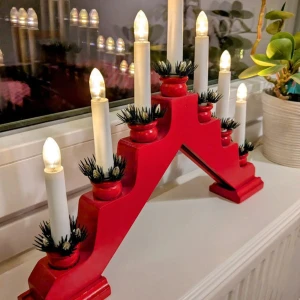 Adventsljusstake - Bilderna visar en röd elektrisk adventsljusstake med sju ljus, dekorerad med gröna plastdetaljer vid ljusbasen. Ljusstaken står i ett fönster och är ansluten med en vit elkabel. Ingen bok syns på bilderna.