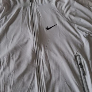 Vit vindjacka med huva från Nike - Säljer en stilren vit vindjacka från Nike med huva och hel dragkedja framtill. Jackan har två fickor med dragkedja och en diskret Nike-logga på bröstet. Perfekt för träning eller när du vill ha en sportig look. Enbart testad och säljs då den inte passar!!