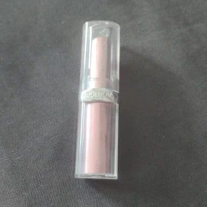 L'Oréal Paris läppstift i nude-rosa nyans - Läppstift från L'Oréal Paris i en mjuk nude-rosa färg. Hylsan är i genomskinlig plast med silverdetalj och själva läppstiftet har en krämig finish. Perfekt för en naturlig look och enkel att ta med i väskan.