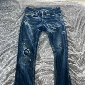 Blå jeans från DonDup med slitningar - Säljer ett par blå jeans från DonDup med tydliga slitningar och hål på flera ställen. Jeansen har klassisk femficksdesign, dragkedja och knapp framtill samt dekorativa sömmar på bakfickan.