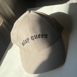 Beige keps med svart text - Snygg beige keps med böjd skärm och svart broderad text 'slay queen' framtill. Justerbar baktill för perfekt passform. Perfekt accessoar för en cool och avslappnad stil.