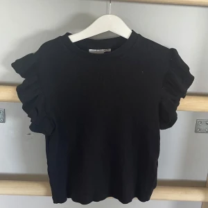 Svart ribbad topp med volangärmar från Gina Tricot - Säljer en svart ribbad topp från Gina Tricot med korta volangärmar. Toppen har rund halsringning och är tillverkad i ett mjukt material som ger en bekväm passform. Perfekt att styla till olika outfits!