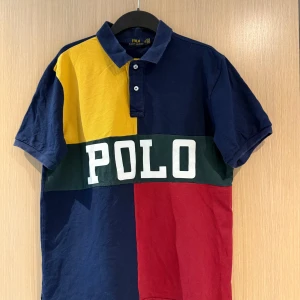 Flerfärgad pikétröja från Polo Ralph Lauren - Säljer en färgstark pikétröja från Polo Ralph Lauren i storlek XL. Tröjan har blockfärger i marinblått, gult och rött med en grön rand och stor vit POLO-text framtill. Klassisk krage och korta ärmar. Perfekt för dig som gillar sportig stil.