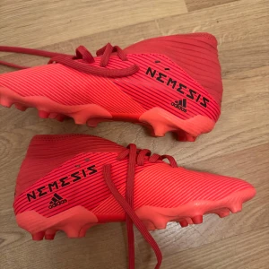 Röda Adidas Nemesis fotbollsskor - Säljer ett par röda Adidas Nemesis fotbollsskor med snörning och låg profil. Skorna har en mönstrad ovandel och är designade för spel på gräs. Perfekta för dig som vill sticka ut på planen!