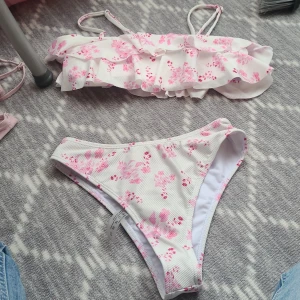 Vit bikini med rosa blommor och volang - Supersöt bikini i vitt med rosa blommigt mönster. Överdelen har smala axelband och bred volang framtill som ger en romantisk vibe. Underdelen är klassisk med samma blommiga print. Materialet är ribbat och känns mjukt mot huden. Perfekt för strandhäng i sommar!