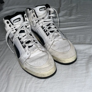 Puma Slipstream vita och svarta sneakers - Puma Slipstream high top sneakers i vitt och svart med klassisk logga på sidan och hälen. Skorna har snörning, rund tå och platt sula. Ovandelen är i skinn och textil, med svart foder och detaljer. Perfekt för dig som gillar retro basketstil. Storlek 46