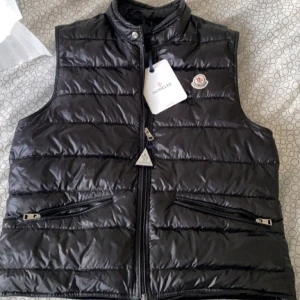 Svart dunväst från Moncler - Snygg svart dunväst från Moncler med glansig finish och klassisk quiltad design. Västen har hög krage, två dragkedjefickor framtill och Moncler-logga på bröstet. Perfekt att slänga över en hoodie eller tröja för extra värme.