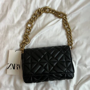 Svart quiltad axelväska från Zara - Snygg svart quiltad axelväska från Zara med chunky guldfärgad kedja som axelrem. Väskan är tillverkad i syntetmaterial och har ett stilrent rutmönster.