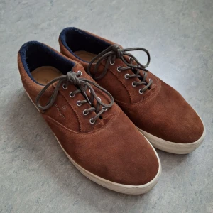 Bruna Gant mockasneakers - Snygga bruna sneakers från Gant i mocka med vita platta sulor och mörkblå foderkant. Skorna har klassisk snörning med grå skosnören och rund tå. Perfekta för en stilren och avslappnad look.