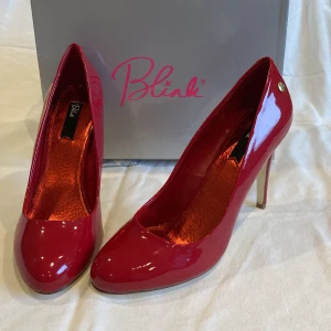Röda lackpumps från Blink, stl 40 - Snygga pumps i djup röd lack från Blink med 11 cm hög klack och rund tå. Skorna har en glansig finish, guldfärgad detalj vid hälen och insida i metallicrött. Tillverkade i syntetmaterial (patent PU). Perfekta för att sticka ut med en färgklick! Oanvända i orginalkartong.