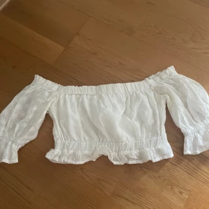 Vit offshoulder blus från Dennis Maglic - Superfin vit offshoulder blus från Dennis Maglic i storlek XS. Blusen har broderade blommor, volangkant nertill och vid ärmslut samt elastiska partier för en snygg passform. Perfekt till sommaren och ger en romantisk vibe.