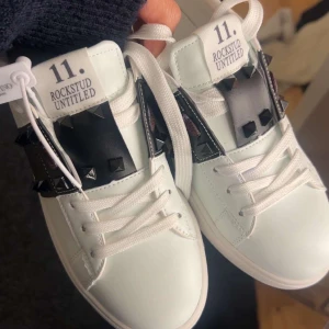 Valentino Rockstud vita sneakers - Snygga helt oanvända vita Valentino Rockstud Untitled sneakers med svarta detaljer och ikoniska nitar. Skorna har snörning och är tillverkade i skinn för en lyxig känsla. Perfekta för dig som vill sticka ut med en edgy och trendig look.