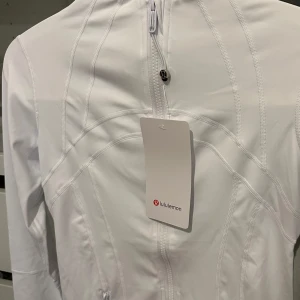 Vit träningsjacka från Lululemon - (PRIS KAN DISKUTERAS VID SNABBAFFÄR) - Slimmad vit träningsjacka från Lululemon med dragkedja framtill, höga fickor med dragkedja och snygga sömdetaljer. Jackan har lång ärm och står upp lite i kragen för en sportig look. Perfekt för dig som gillar stilrena träningskläder. 