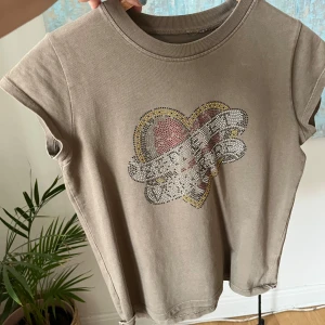 Beige t-shirt med hjärtmotiv Zadig & Voltaire - Cool beigegrön t-shirt från Zadig & Voltaire med korta ärmar och ett stort hjärtmotiv framtill i glittriga nitar i rött, gult, svart och silver. Skön passform och tillverkad i mjuk bomull. Perfekt för dig som gillar edgy detaljer och statement-plagg.