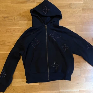 Svart hoodie  - Cool svart hoodie med hel dragkedja och huva. Tröjan har flera stora korsliknande mönster i färgglada stenar på både fram- och baksidan samt ärmarna. Perfekt för dig som vill sticka ut med en unik streetstyle-look.