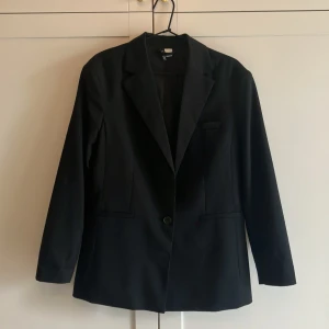 Svart kavaj från H&M - Klassisk svart kavaj från H&M med enkelknäppning och smal krage. Rak passform med två fickor framtill och en bröstficka. Perfekt för en stilren look och lätt att matcha med olika outfits. Oversize modell. Storlek XS