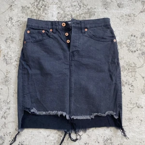 Svart jeanskjol med fransad kant 157 - Svart jeanskjol från 157 med hög midja, knappar framtill och fransad nederkant. I storlek S. 