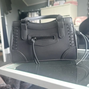 Grå handväska från Gina Tricot - Stilren svart handväska från Gina Tricot i syntetmaterial. Väskan har ett coolt flätat detaljmönster på sidorna, ett litet ytterfack med dragkedja och dubbla handtag. Perfekt storlek för det viktigaste och med snygga sömmar som ger extra edge.