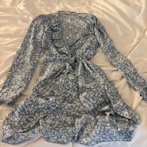 Blommig omlottklänning från ONLY - Supersöt blå och vit omlottklänning från ONLY med blommigt mönster. Klänningen har långa ärmar, volangdetaljer och knyt i midjan. Perfekt för dig som gillar en feminin och fräsch stil. Lätt och skönt material, perfekt för varmare dagar.