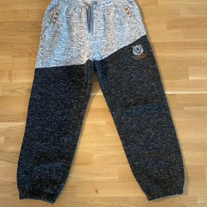 Gråmelerade mjukisbyxor med emblem - Snygga mjukisbyxor i gråmelerad och mörkgrå färg med broderat emblem på benet. Byxorna har elastisk midja med snörning, muddar vid benslut och dekorativa knappar vid fickorna. Perfekta för chill eller träning.