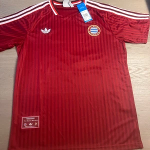 Bayern München Musiala 42 Adidas tröja - Röd kortärmad Bayern München fotbollströja från Adidas med Musiala och nummer 42 på ryggen. Tröjan har diskreta vertikala ränder, klubbmärke på bröstet och klassiska vita Adidas-ränder på axlarna. Tillverkad i lätt och ventilerande material.