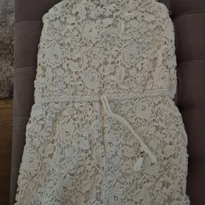 Vit jumpsuit från Vintage Stories - Superfin jumpsuit från Vintage Stories med vackert blommigt spetsmönster och knytband i midjan. Jumpsuit har smala axelband och är perfekt för sommarens alla tillfällen. Den är fodrad och känns både romantisk och trendig.