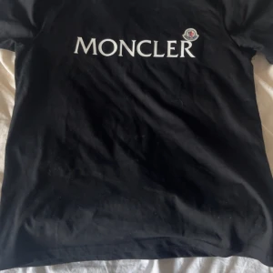 Svart Moncler t-shirt med logga - Svart t-shirt från Moncler med stor vit logotyp på bröstet och klassisk rund hals. T-shirten har korta ärmar och är gjord i mjuk bomull. En clean och stilren tee med exklusiv känsla och broderad Moncler-patch på ärmen.