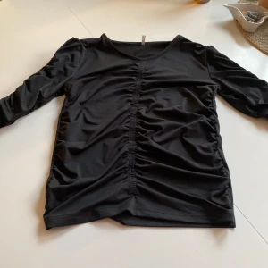 Svart långärmad topp från Only - Svart långärmad topp från Only med snygg rynkdetalj längs hela framsidan och på ärmarna. Tunt och stretchigt material som känns skönt mot huden. Perfekt för dig som gillar en stilren men ändå trendig look.