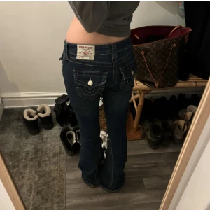 True Religion bootcut jeans mörkblå - Mörkblå jeans från True Religion med bootcut passform och låg midja. Snygga kontrastsömmar, stora fickor bak med dekorativa knappar och klassisk logga på bakfickan. Jeansen har en klassisk femficksdesign och är tillverkade i stretchig denim.