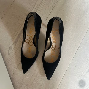 Svarta pumps från Sam Edelman - Klassiska svarta pumps från Sam Edelman med spetsig tå och hög klack. Skorna är i mocka med snygga detaljer i skinn vid hälen. Perfekta för dig som vill ha en stilren och tidlös look.
