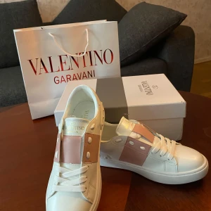 Valentino Garavani Open sneakers vit/rosa - Snygga Valentino Garavani Open sneakers i vitt skinn med bred rosa rem över vristen och ikoniska nitar på hälen. Platt sula med mönstrad undersida och klassisk snörning. Perfekta för dig som vill ha en lyxig och trendig look.